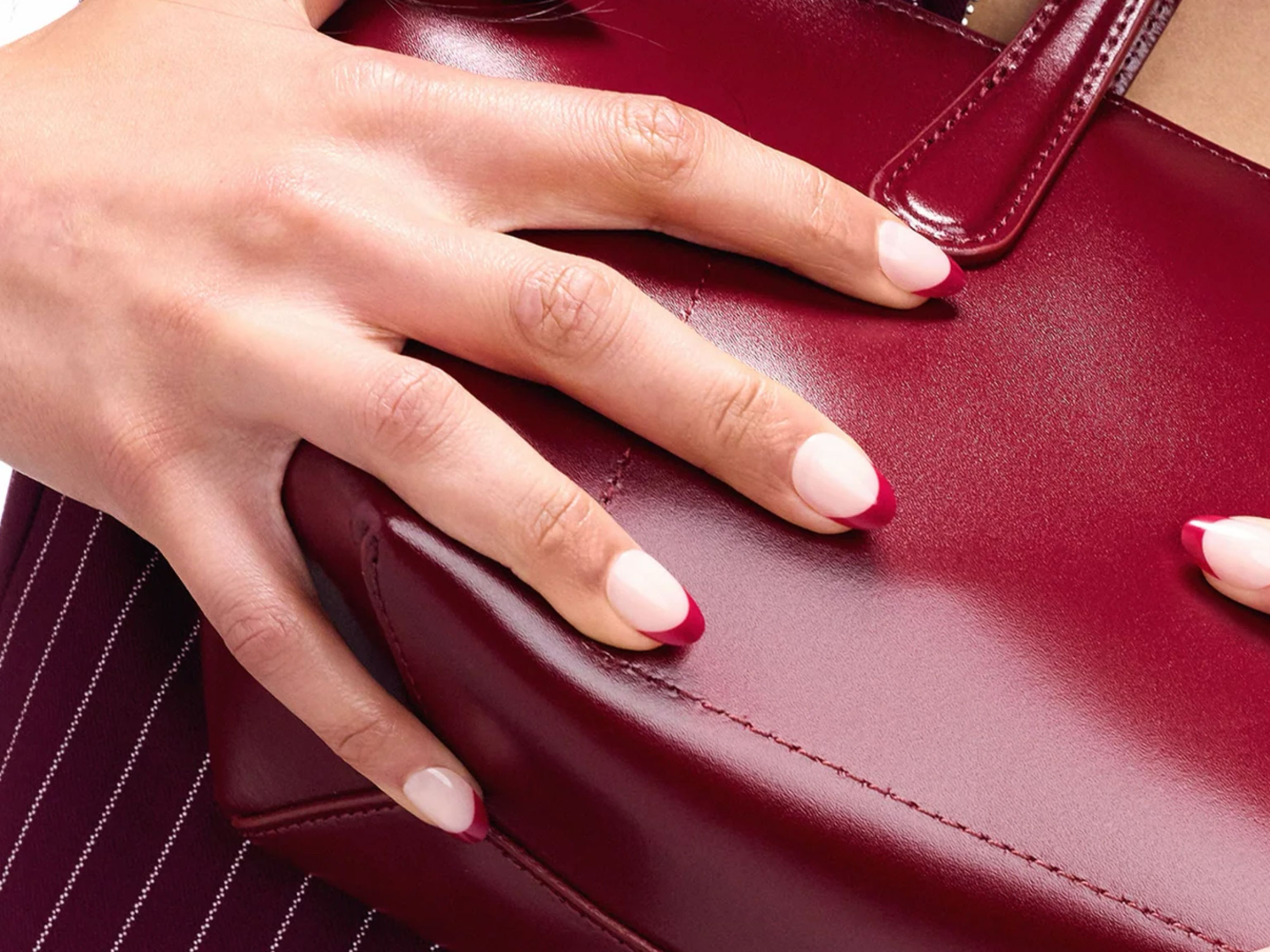 Close-up of a hand with blush pink press-on nails featuring deep red French tips, resting on a burgundy leather handbag
